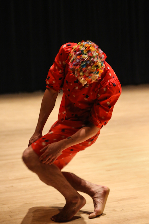 Brian Strimpel in a red house dress and mask. He squats down as he balances on the balls of his feet.